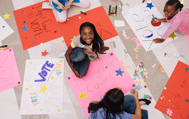 Overhead view of four girls creating posters that encourage people to vote. One Cadette is looking up at the camera.