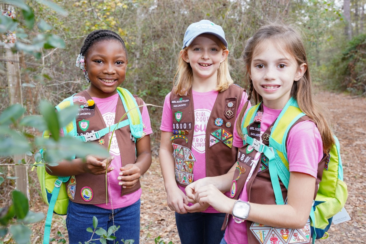 Three Girl Scouts wearing Brownie vests and backpacks, standing in wooded area